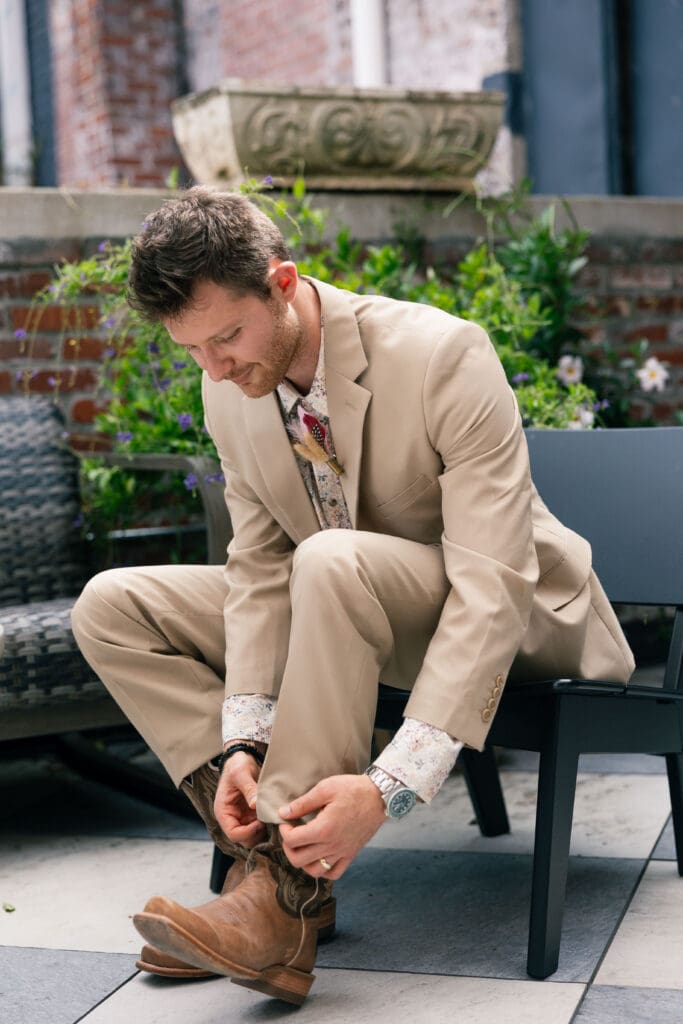 Groom in a tan suit ties his brown boot while seated in a garden courtyard, highlighting why comfortable getting ready spaces matter when comparing Charleston SC wedding venues.