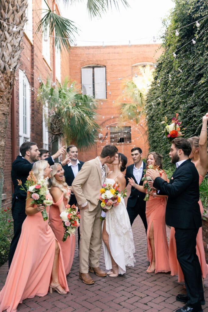 Wedding party gathers around the newlyweds on a brick courtyard with colorful florals and joyful expressions, reflecting candid Charleston wedding photography.