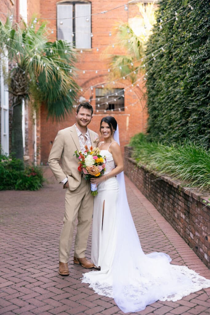 Bride and groom pose along a brick courtyard framed by greenery and historic architecture, reflecting the visual character of unique Charleston SC wedding venues.