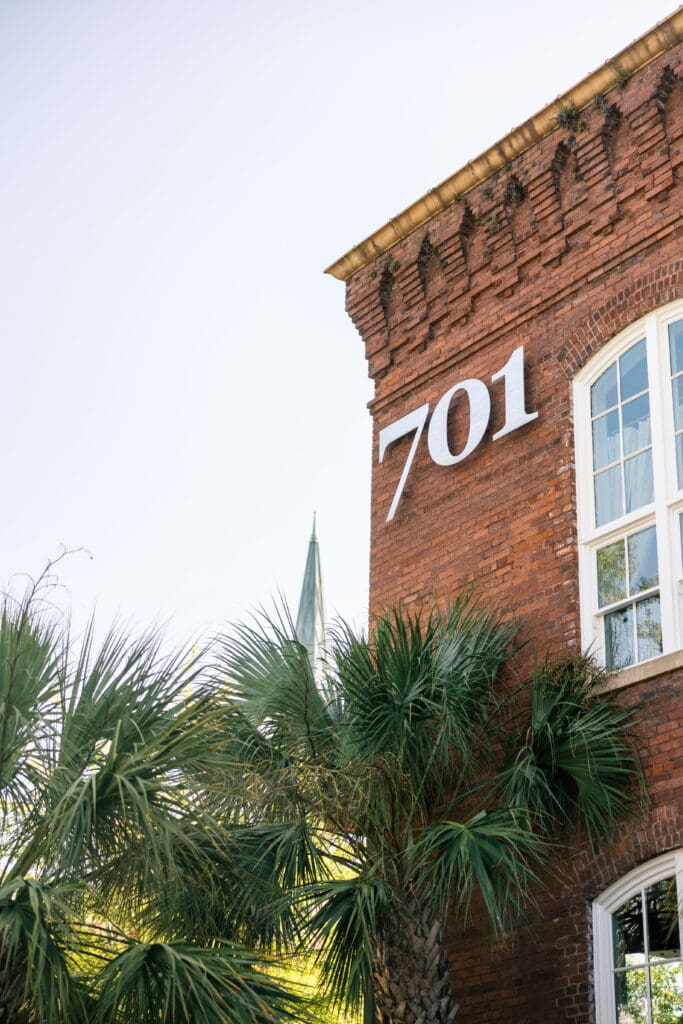 Historic red brick exterior marked with the 701 address rises above palmettos, showcasing the architectural character of the 701 Whaley wedding venue in Columbia.