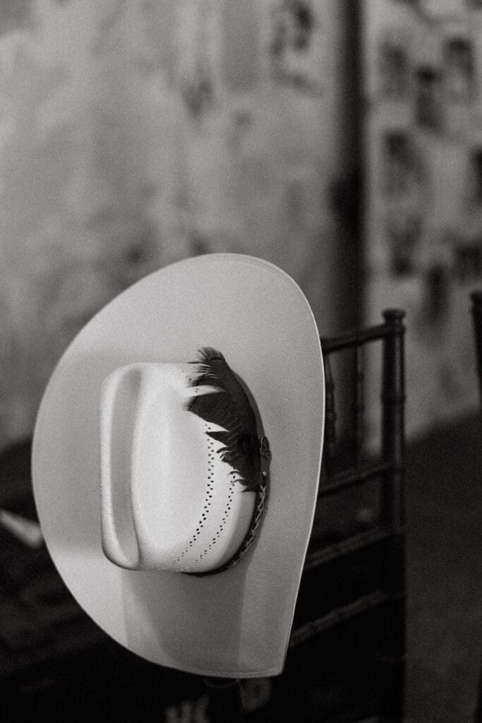 Black-and-white photograph of a white cowboy hat with a decorative feather resting on a chair