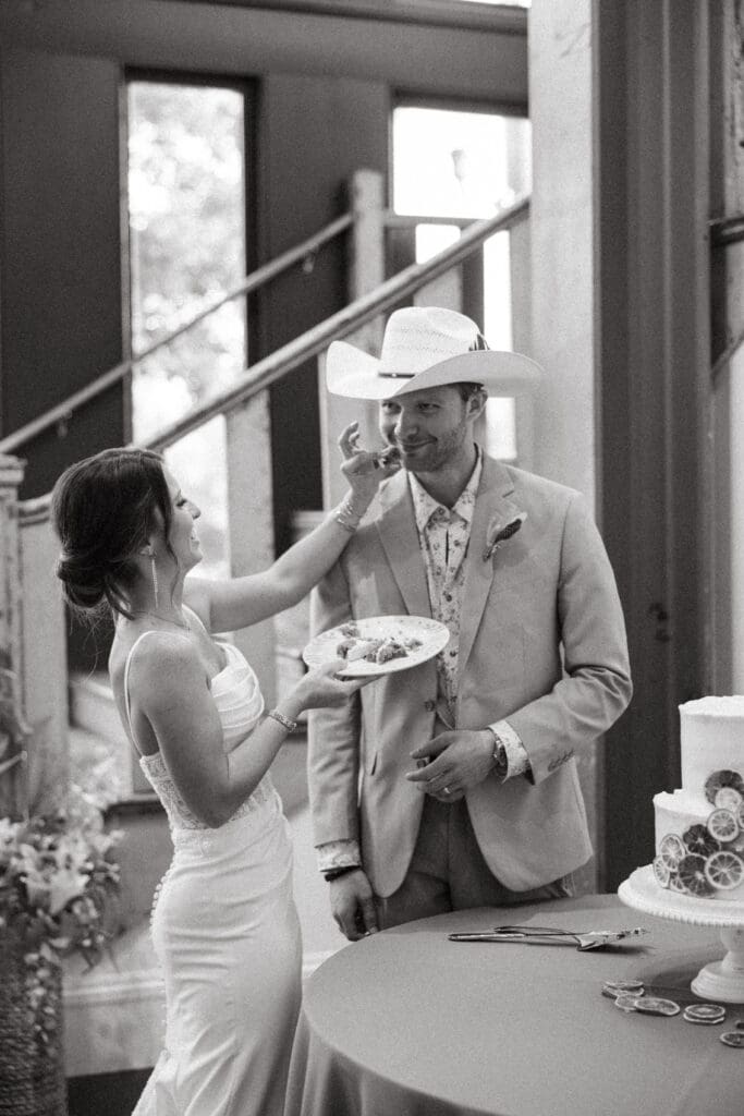 Bride playfully feeds the groom during their reception in a candid black and white photograph, capturing the relaxed spirit of a wedding at 701 Whaley.