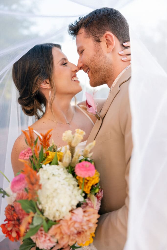 Bride and groom smiling beneath a veil while holding a colorful wedding bouquet