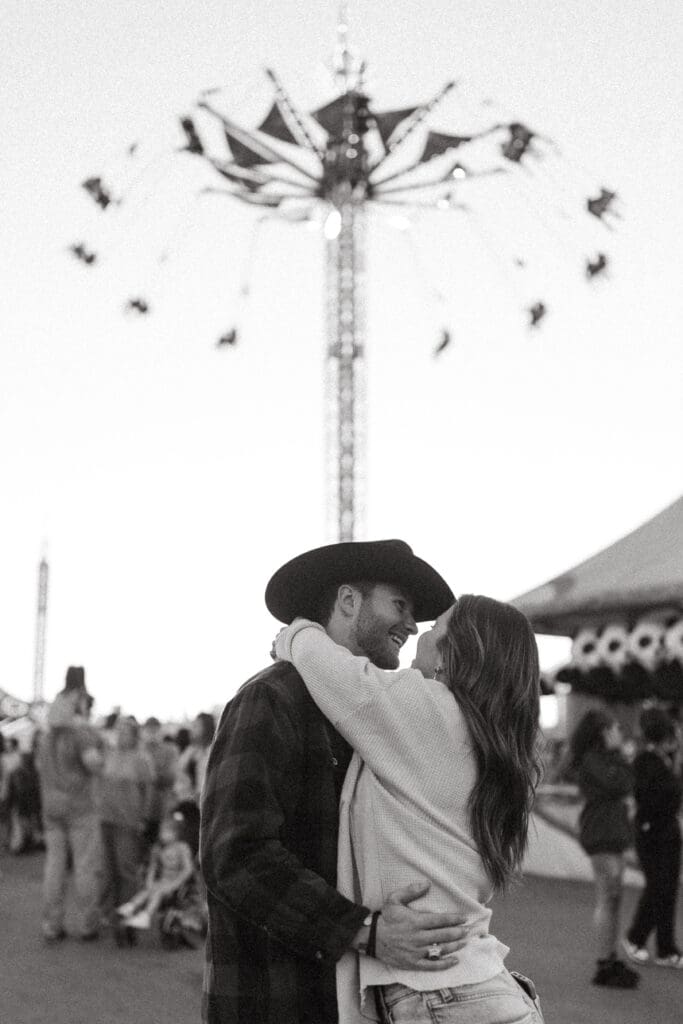 Couple embraces beneath a carnival ride in a romantic black and white portrait, blending genuine affection with candid Charleston SC wedding photography.
