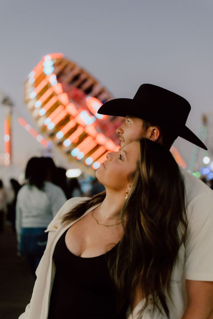 Woman smiles beneath a black cowboy hat with colorful carnival lights glowing behind her, part of a relaxed and personality driven Charleston couples session.