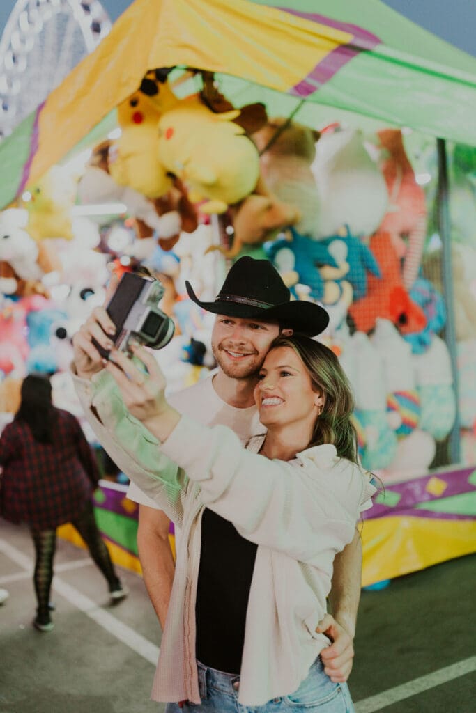 Smiling couple takes a selfie together against a vibrant carnival backdrop, showing the fun and unscripted side of a Charleston engagement photo session.