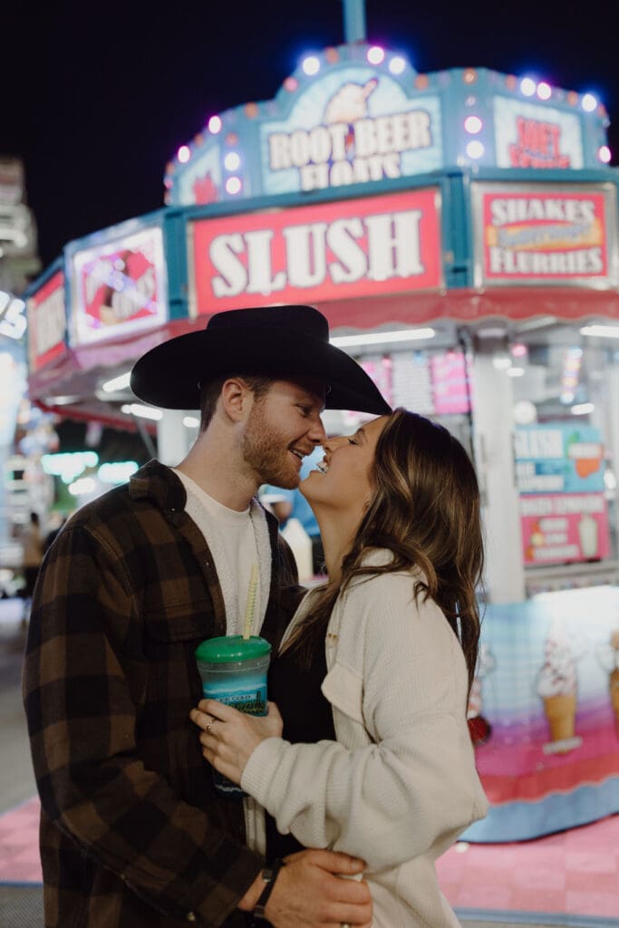 Couple in casual outfits leans in for a kiss beneath colorful carnival lights, bringing a playful nighttime mood to unique Charleston engagement photos.