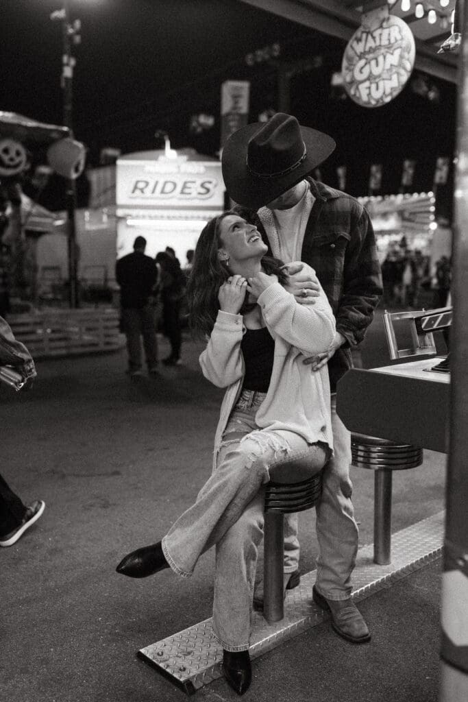 Black and white photograph of a couple sharing an affectionate moment at a carnival game, finishing their romantic Charleston engagement session with a candid, documentary feel.