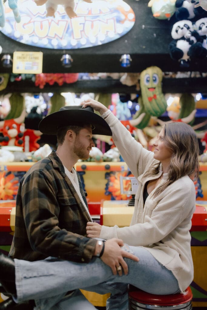 Playful couple sits together at an arcade game as the bride to be tips her partner's hat, a colorful and candid Charleston engagement session moment.
