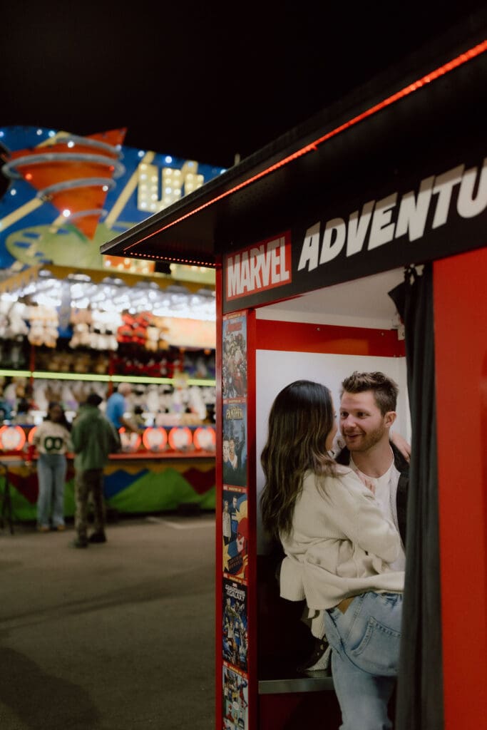 Couple peeks around the entrance of a brightly lit carnival attraction, creating a playful composition for couples seeking creative engagement photo ideas in Charleston.