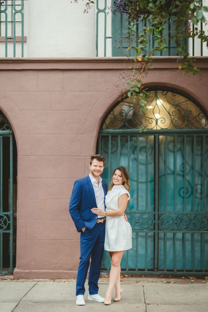 Couple poses against a blush colored historic facade with arched ironwork, showcasing the architectural charm of a wedding venue in downtown Charleston SC.