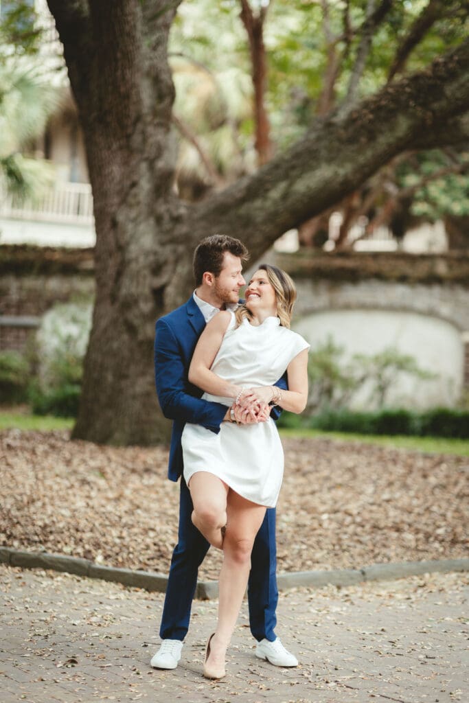 Couple embraces beneath the sweeping branches of a mature tree, creating a romantic portrait inspired by the timeless scenery of a Charleston engagement session.