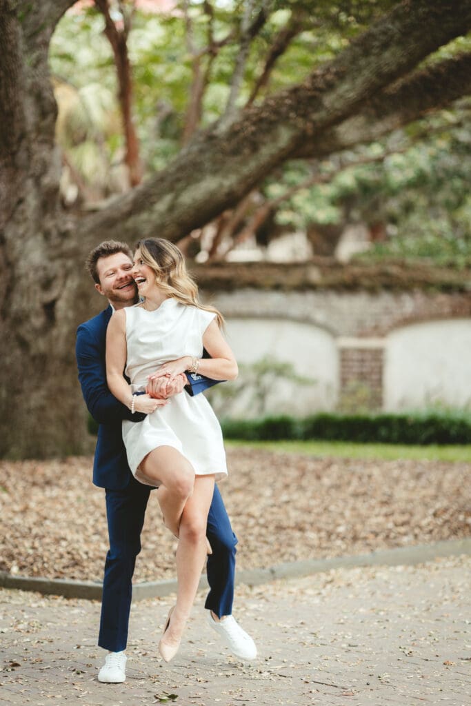 Groom lifts his bride beneath sprawling tree branches as they laugh together, a relaxed moment suited to romantic Charleston wedding photography.