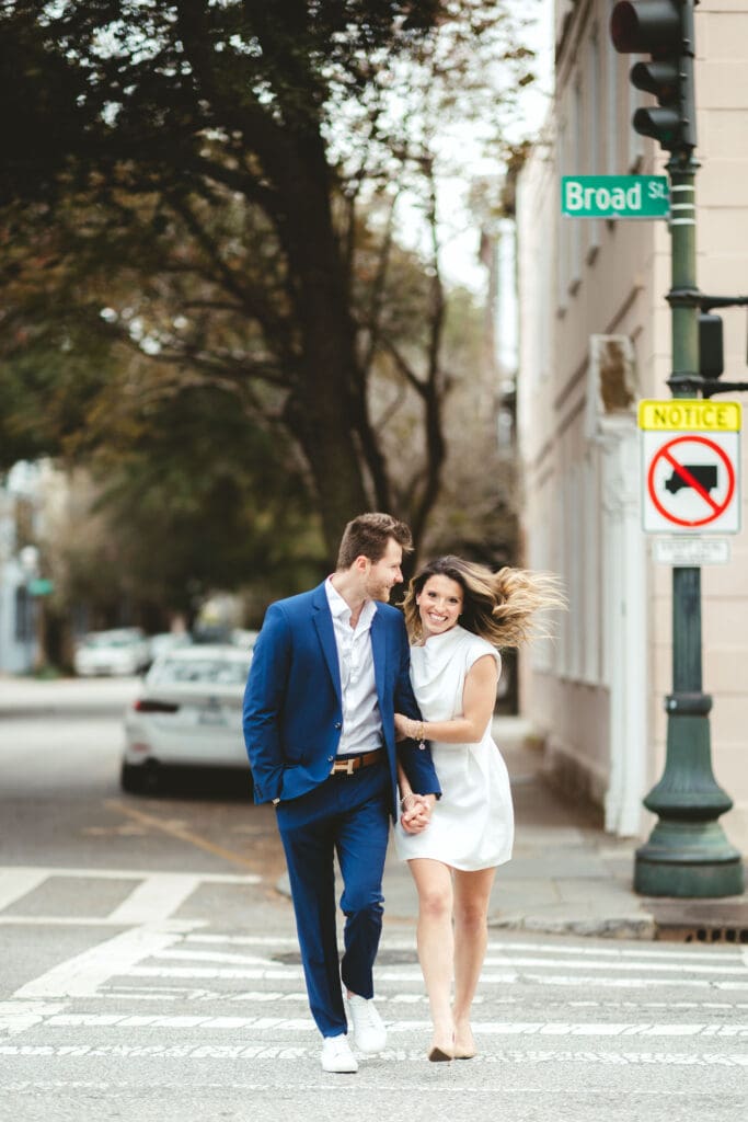 Couple walks hand in hand near the Broad Street sign beneath mature trees and historic architecture, capturing the relaxed charm of a downtown Charleston SC engagement session.