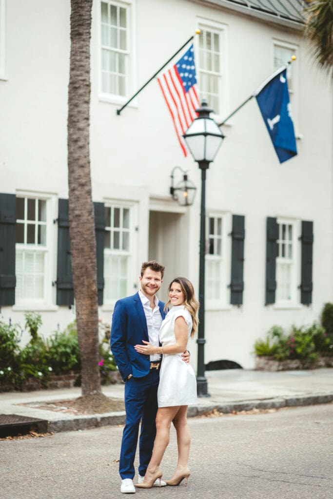 Couple poses outside a classic white Charleston building with palm trees and flags, highlighting the architectural character found throughout historic downtown Charleston.