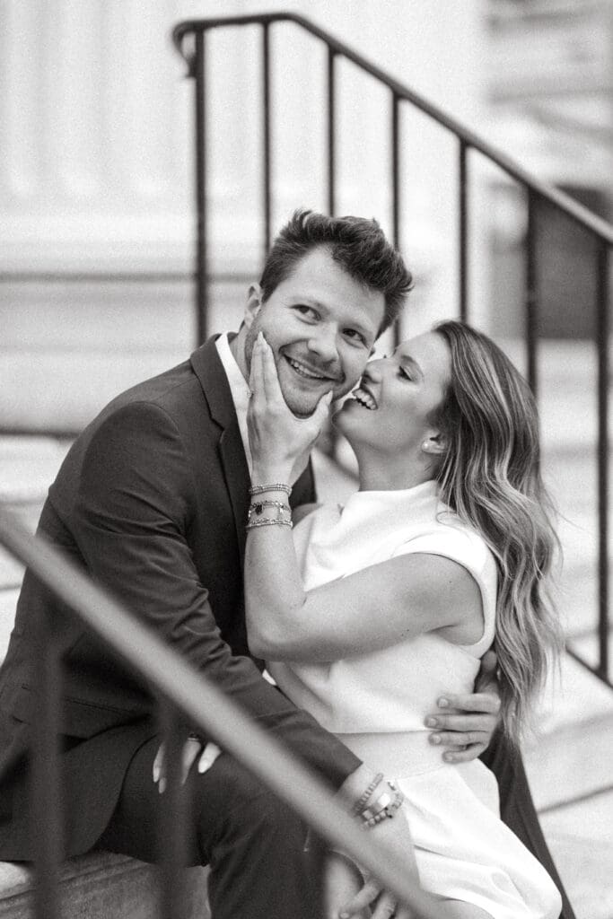 Woman kisses her partner's cheek as they embrace beside a staircase, captured in timeless black and white Charleston wedding photography.
