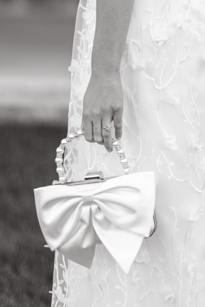 Close up of a bride carrying a structured white bow handbag against her textured wedding dress, an editorial detail from an elegant Charleston wedding celebration.