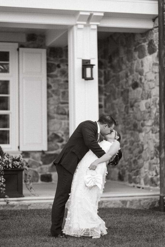 Black and white portrait of a groom dipping and kissing his bride outside a stone and white columned building, capturing the romantic character couples seek in Charleston SC wedding venues.