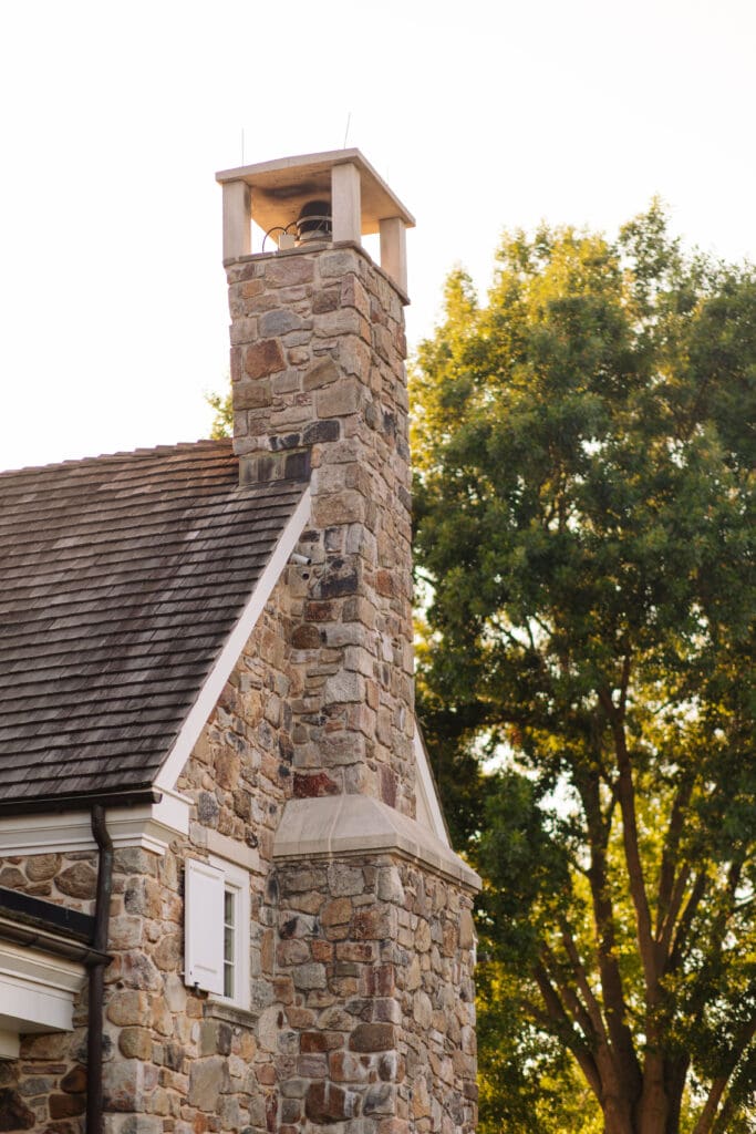 Sunlight filters through mature trees behind a stone cottage and chimney, capturing the architectural character that makes Lowcountry wedding venues distinctive.