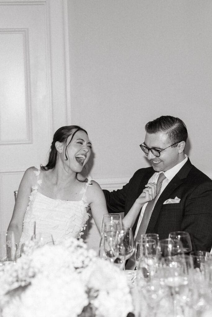 Newlyweds laugh together at their reception table surrounded by glassware and floral details, captured through natural Charleston wedding reception photography.