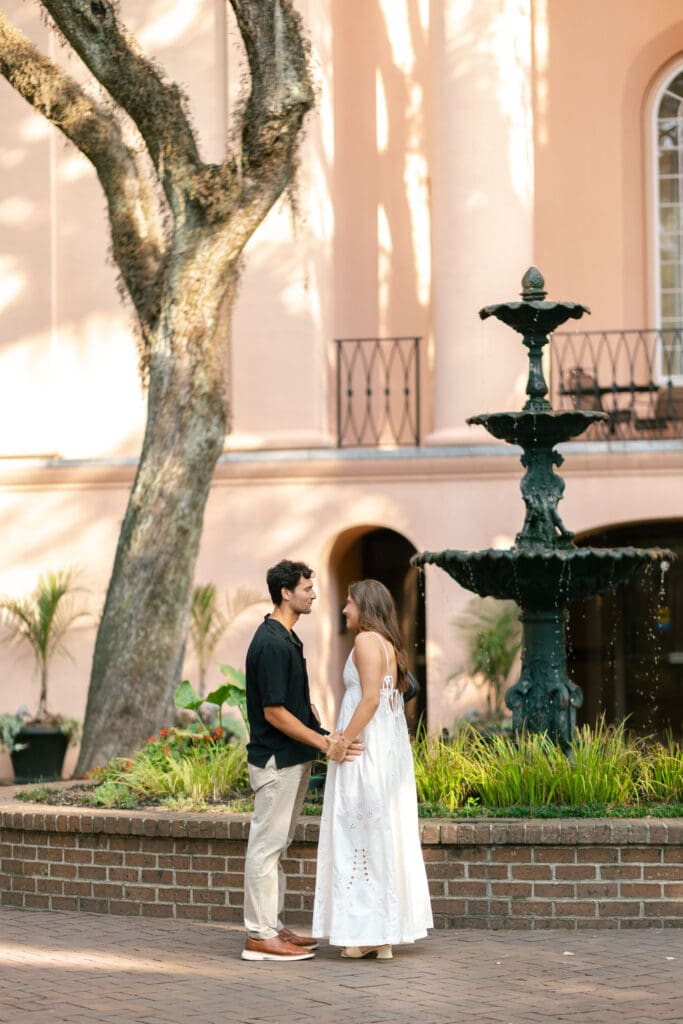 Newly engaged couple holds hands beneath mature trees beside a fountain and pastel Charleston architecture, a romantic setting among the best places to propose in Charleston SC.