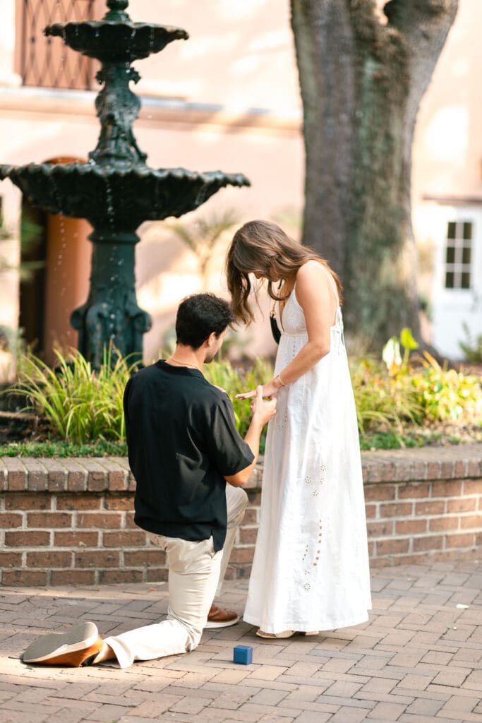 A man kneels with a ring as his partner reaches toward him beside a brick garden and fountain, capturing the emotion of a surprise proposal in Charleston SC.