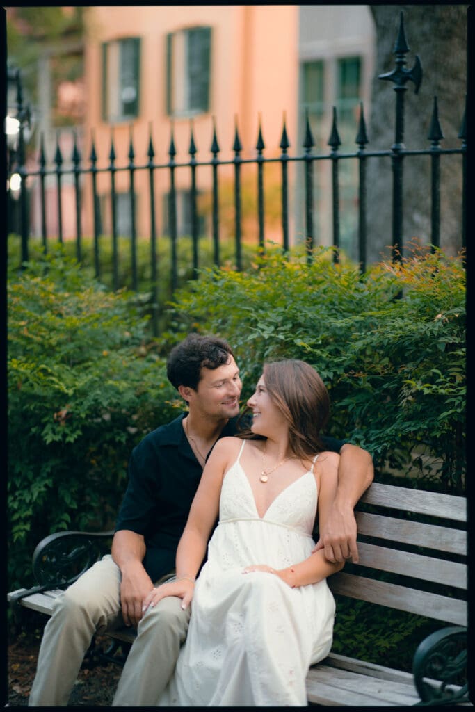 Relaxed couple sits close together on a garden bench framed by greenery and wrought iron, capturing the quiet charm of a downtown Charleston engagement session.