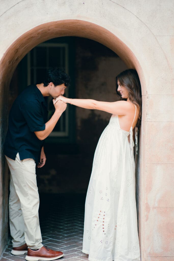 Man gently kisses his partner’s hand beneath a rounded architectural archway, adding an editorial touch to their Charleston SC engagement photos.