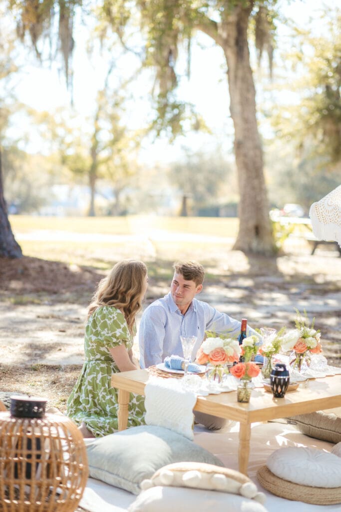 Couple sits together at an intimate outdoor table beneath trees near the water, a relaxed setting to consider when exploring small wedding venues Charleston SC.