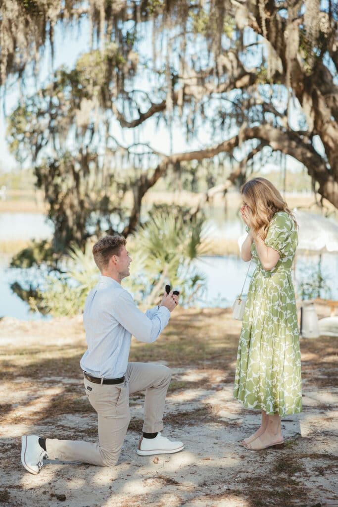 Man proposes beneath a sprawling oak draped in Spanish moss as his surprised partner looks down at him, an intimate Lowcountry proposal location in Charleston.