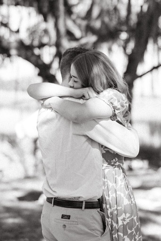 Black and white portrait captures the newly engaged couple in a joyful embrace beneath moss covered trees, preserving the emotion of a Charleston proposal.