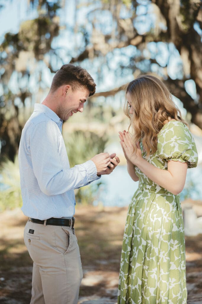 A woman admires her new engagement ring while her fiancé holds her hand beneath moss covered branches, a candid moment from a Charleston proposal photography session.