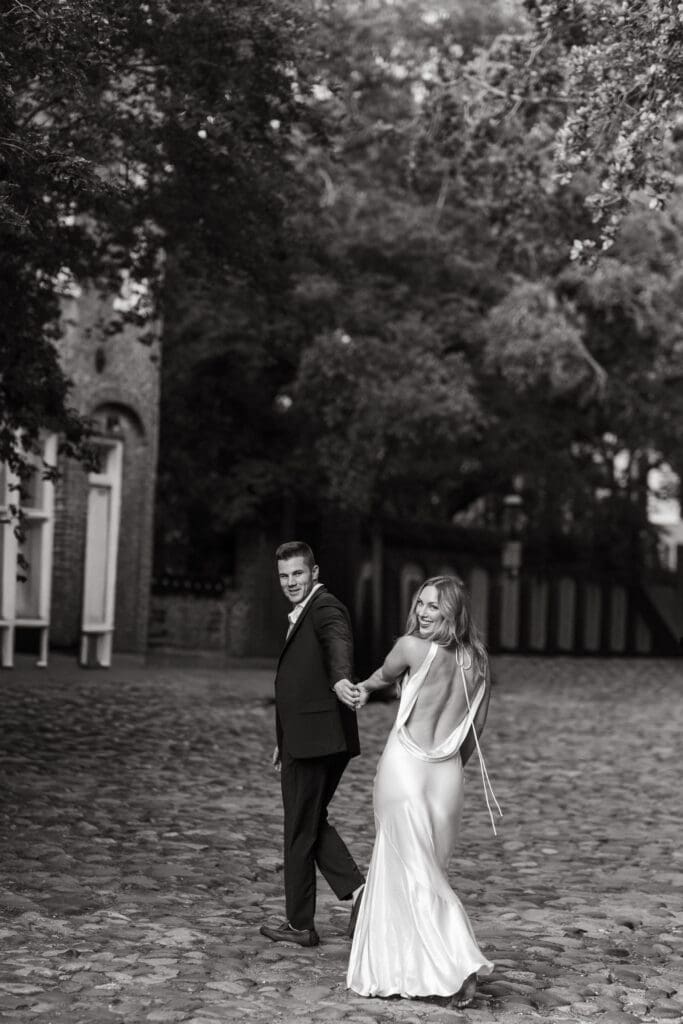 Elegantly dressed couple walks hand in hand along a cobblestone street framed by trees, showcasing the historic atmosphere of engagement photos in downtown Charleston SC.