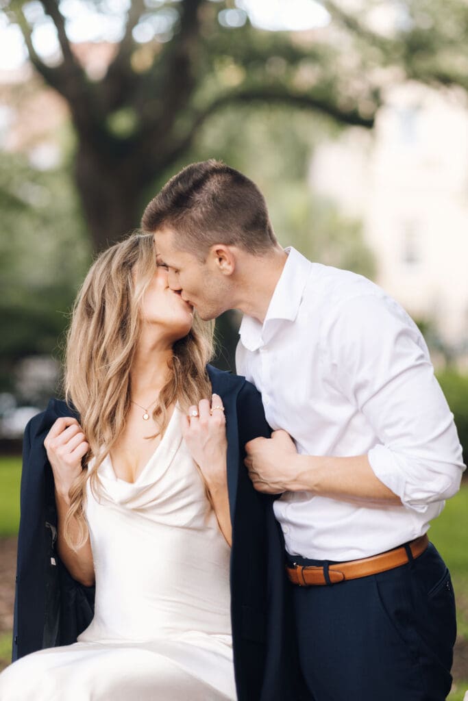 Couple shares a soft kiss beneath leafy trees while dressed in polished neutral tones, a candid moment suited to romantic Charleston engagement photography.
