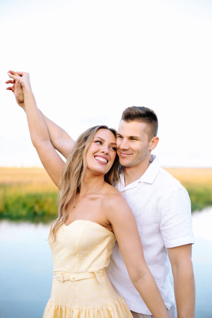 Smiling woman in a pale yellow dress raises her arm as her partner stands close behind her by the waterfront, a joyful ending to their Charleston SC engagement photography.