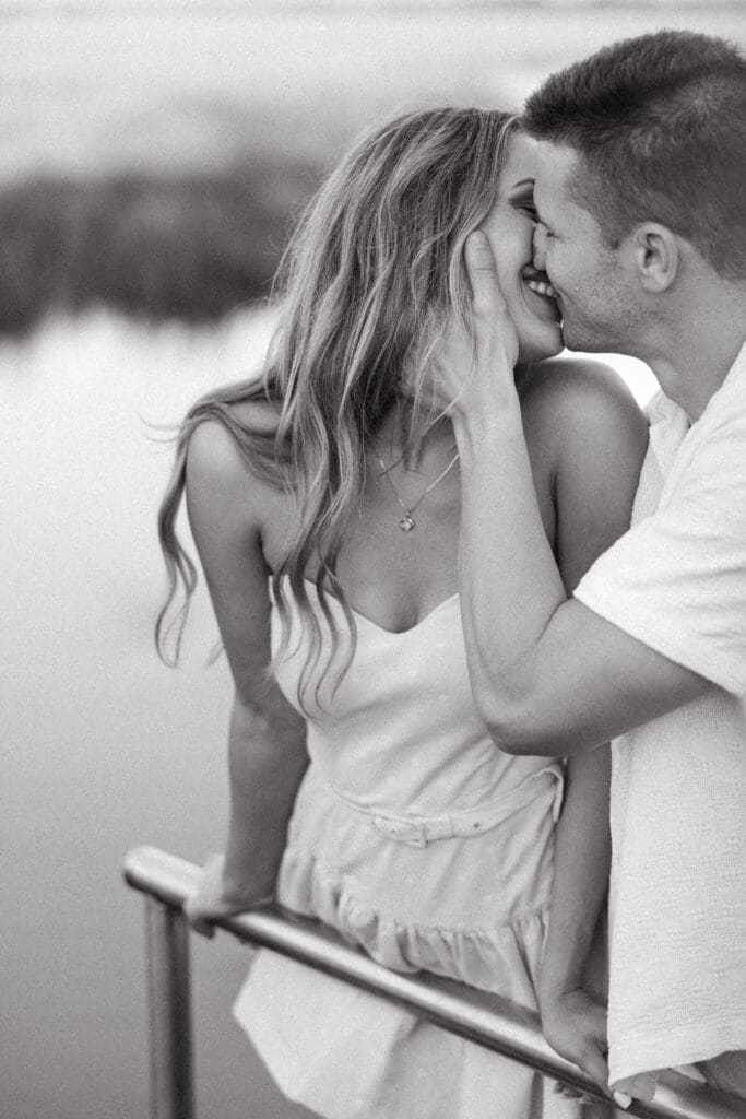 Black and white portrait of an engaged couple sharing a kiss beside the water, reflecting the candid style of a Charleston engagement photographer.