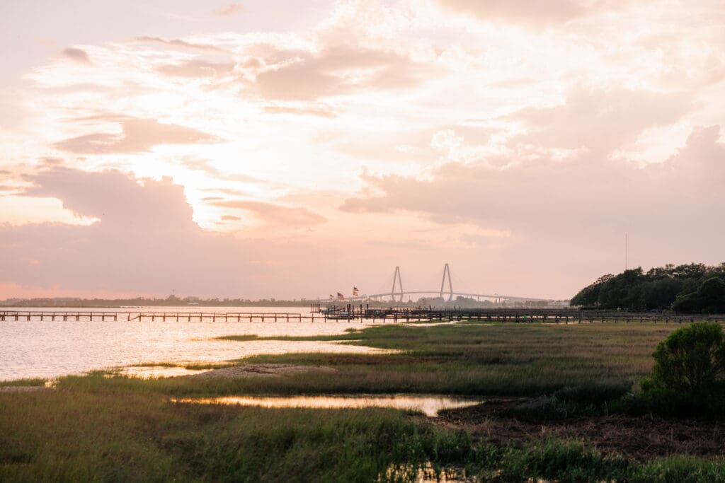 Sunset settles over a Lowcountry marsh with the Charleston skyline and bridge in the distance, a scenic perspective on wedding venues on the water in Charleston SC.