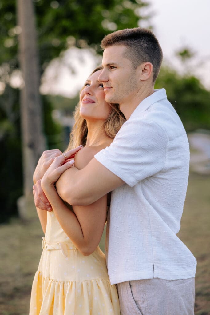 Man embraces his fiancée from behind as they smile together outdoors, creating a warm and natural portrait during a Charleston SC engagement session.