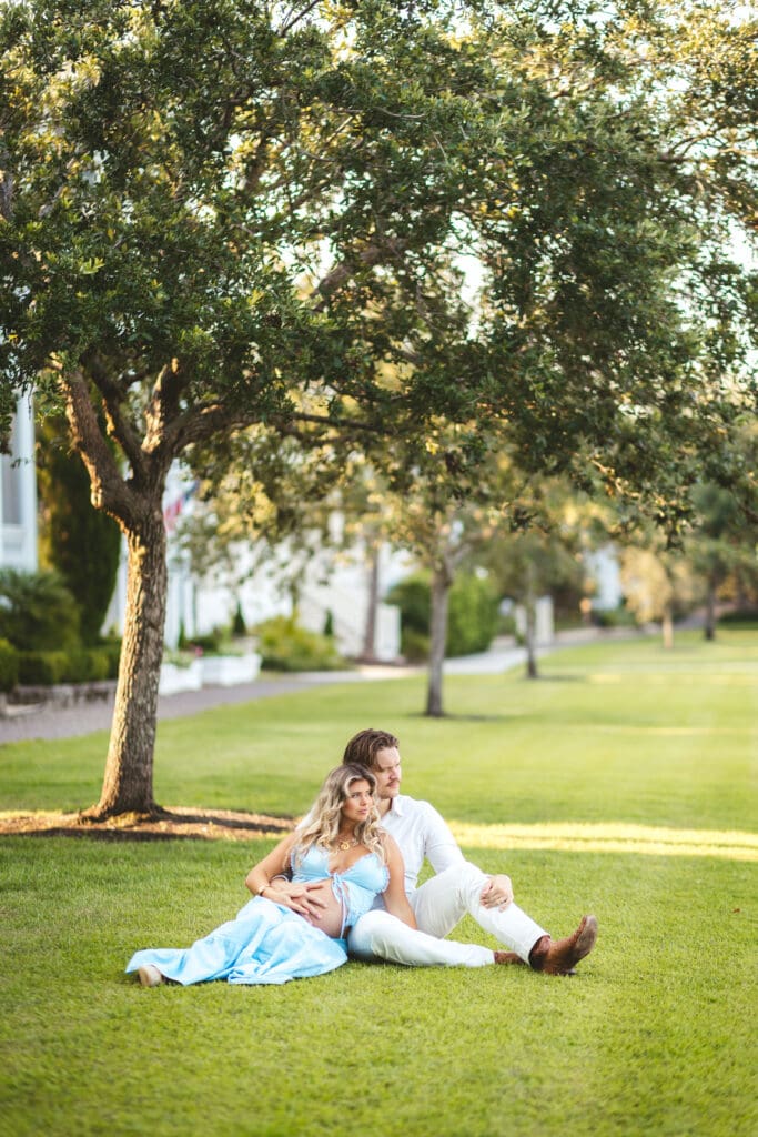 Expectant parents relax together on a sunlit lawn beneath a leafy tree, showcasing the soft outdoor scenery found around I’On in Mount Pleasant.