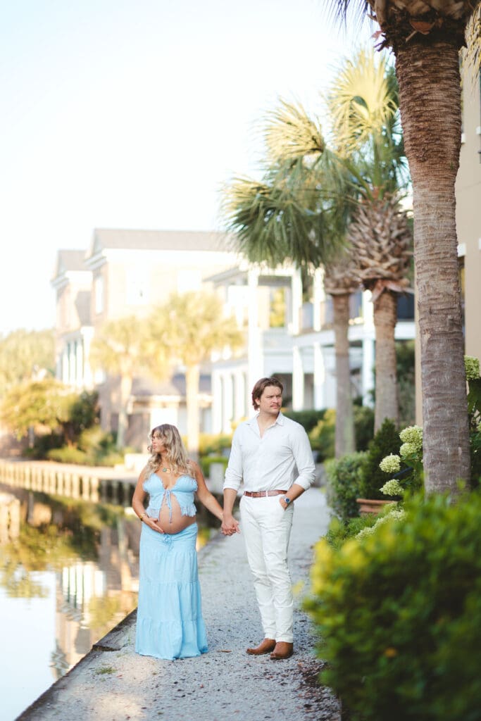 Couple strolls beside the water and palm lined homes in coordinating light tones, reflecting the coastal elegance surrounding a Creek Club I’On wedding setting.