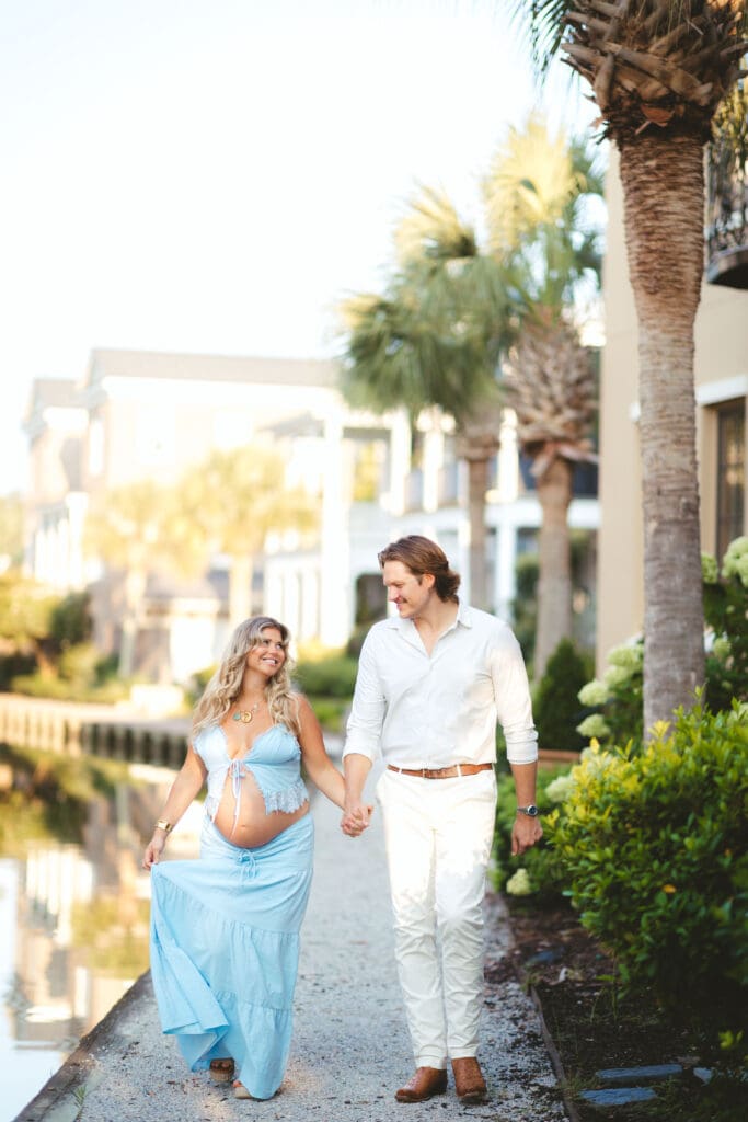 Expectant couple strolls along a palm lined waterfront path, showcasing the relaxed coastal atmosphere associated with Charleston SC waterfront wedding venues.