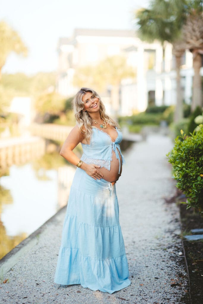 Expectant mother in a flowing blue dress poses beside the water in warm natural light, highlighting the waterfront character of I’On in Mount Pleasant SC.
