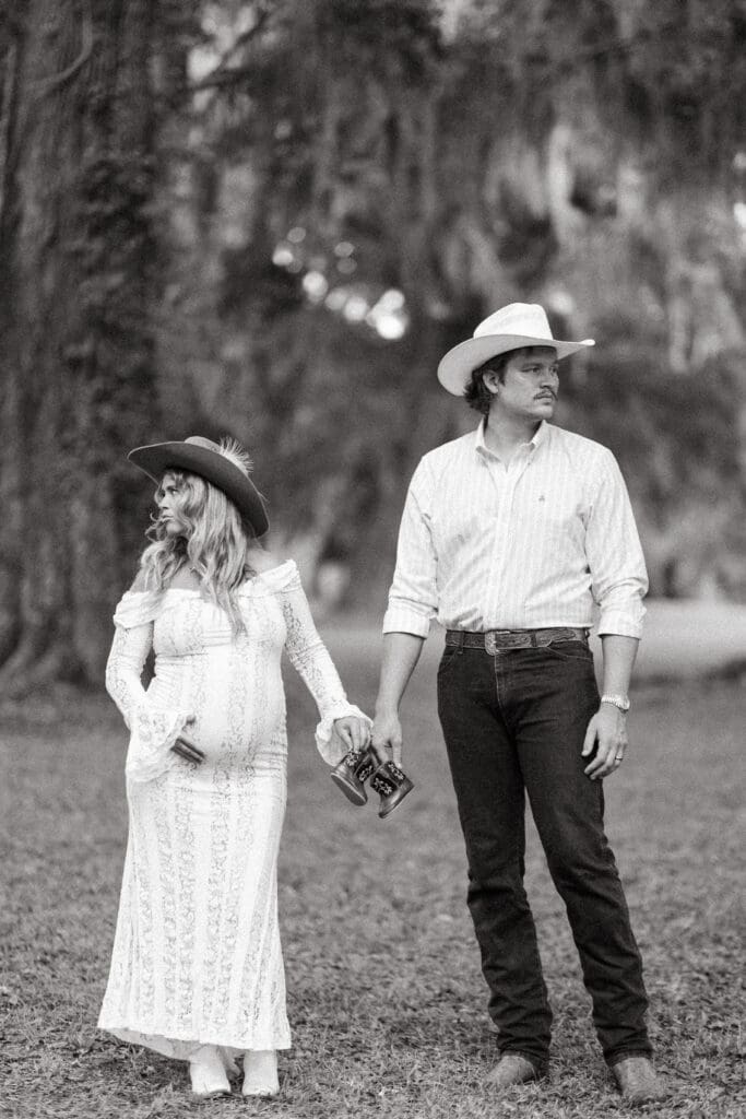 Expectant couple walks hand in hand beneath moss draped trees, creating a timeless black and white portrait inspired by the Lowcountry setting around Creek Club at I’On.