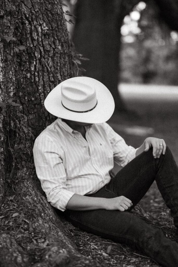 Man in a wide brim hat rests against a large tree in a quiet black and white portrait, capturing the relaxed Lowcountry style of Mount Pleasant photography.